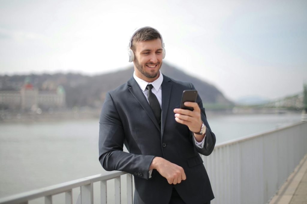 Smiling businessman in a suit listens to music on smartphone by a river.